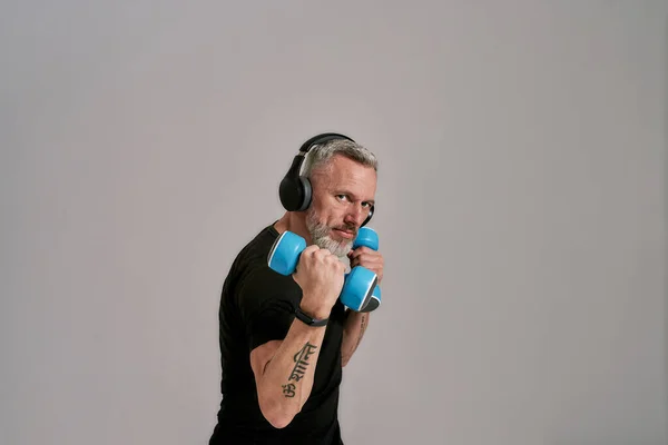 Uomo muscoloso di mezza età in t-shirt nera e cuffie guardando la fotocamera che tiene i manubri blu, in posa in studio su sfondo grigio — Foto Stock
