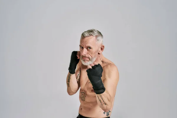 Mezza nuda uomo atletico di mezza età, kickboxer guardando la fotocamera durante la boxe, praticare pugni in studio su sfondo grigio — Foto Stock