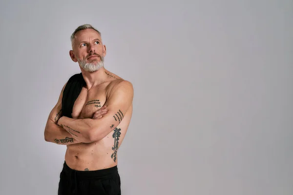 Medelålders muskulös man tog av svart t-shirt, visar sin kropp, tittar åt sidan medan poserar i studio över grå bakgrund — Stockfoto