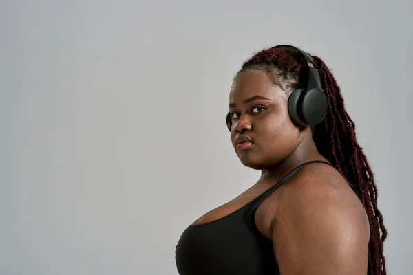 Plumpe, plus Size afrikanische Amerikanerin in Sportbekleidung und Kopfhörer, die in die Kamera schaut, im Studio vor grauem Hintergrund stehend. Konzept des Sports, gesunder Lebensstil, positiver Körper, Gleichberechtigung — Stockfoto