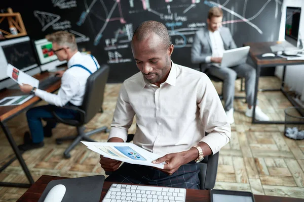 Imagina más. Comerciante africano enfocado sentado junto al escritorio y estudiando informes analíticos mirando a través de papeles en la oficina. Sus colegas están trabajando en segundo plano. — Foto de Stock