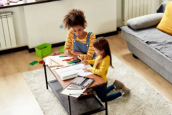Dedicated. Caucasian little girl spending time with african american baby sitter. They are drawing, learning to write letters, sitting on the floor — Stock Photo, Image