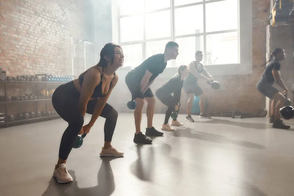 Concentra-te no Fitness. Tiro de comprimento total de jovem mulher atlética alegre levantando um peso ao ter treino no ginásio industrial. Formação em grupo, conceito de trabalho em equipa — Fotografia de Stock