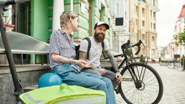 Two cheerful couriers, young man and woman sitting on the bench and talking outdoors while delivering food and products, using scooter and bike — Stock Photo, Image