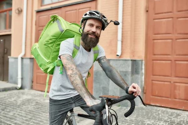 Idealna dostawa. Brodaty dostawca w kasku z torbą termiczną lub plecakiem uśmiechający się do kamery, jeżdżący na rowerze po mieście, dostarczający jedzenie. Kurier, koncepcja usługi dostawy — Zdjęcie stockowe