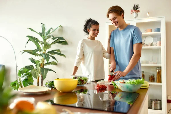 Giovane uomo che taglia verdure mentre la donna lo guarda. I vegetariani che preparano pasti sani in cucina insieme. Vegetarismo, concetto di cibo sano — Foto Stock