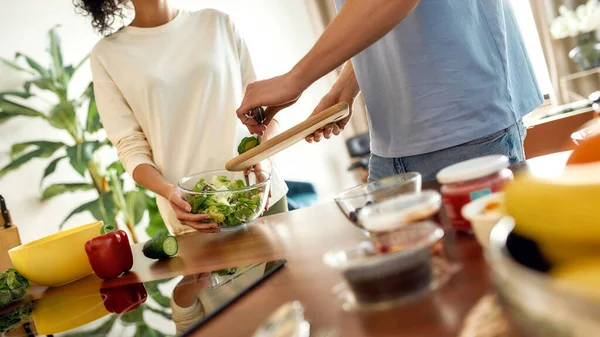男人把切碎的黄瓜放进盘子里，女人帮他，拿着沙拉盘。素食主义者在厨房里一起准备健康的饭菜 — 图库照片