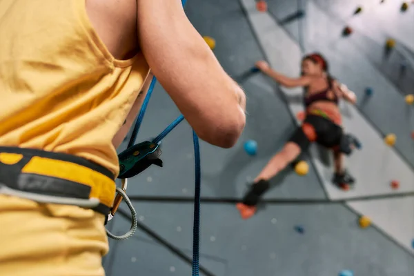 Ausgeschnittene Ansicht eines männlichen Instruktors mit Seil, während die Frau in Sicherheitsausrüstung und Gurtzeug an der künstlichen Kletterwand trainiert — Stockfoto