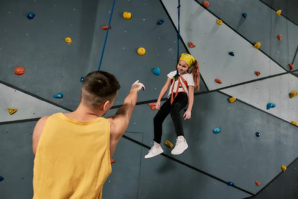 Männlicher Instruktor hält Seil, spricht mit einem Mädchen in Sicherheitsausrüstung und Gurtzeug, während sie an der künstlichen Kletterwand trainiert — Stockfoto