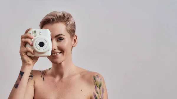 Snap. Retrato de uma bela mulher tatuada com nariz perfurado e cabelo curto usando câmera de foto instantânea, de pé isolado sobre fundo cinza — Fotografia de Stock