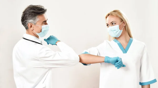Coronavirus megelőzés. Férfi és női orvosok, akik védőmaszkot viselnek, könyökökkel üdvözlik egymást, szürke háttér előtt állnak. — Stock Fotó