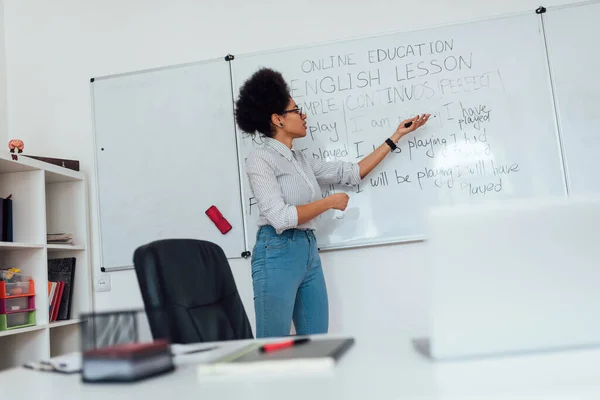 A dar aulas online. Jovem afro-americana professora em roupas casuais perto do quadro branco e explicando a gramática inglesa — Fotografia de Stock