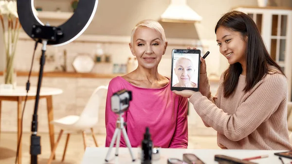 전 보다 낫지. 젊은 여성 블로거, 메이크업 아티스트 태블릿 pc 를 사용하여 화장을 한 후 중년 여성의 얼굴 앞에 들고 있는 모습 — 스톡 사진