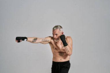 Yarı çıplak orta yaşlı atletik adam, kickboksçu boks yaparken yana bakıyor, gri arka plan üzerinde stüdyoda yumruk pratiği yapıyor.