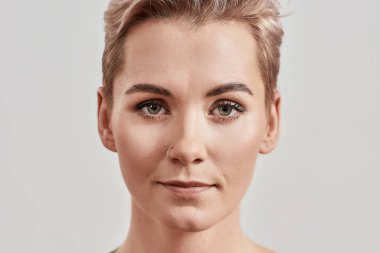 Kısa saçlı, mükemmel ciltli ve makyajlı çekici bir kadının açık arkaplanda izole edilmiş kameraya bakarken çekilmiş yüzü.