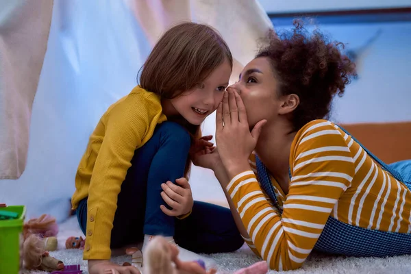 African american woman baby sitter entertaining caucasian cute little girl. They are gossiping and telling secrets sitting in kids room — Stock Photo, Image