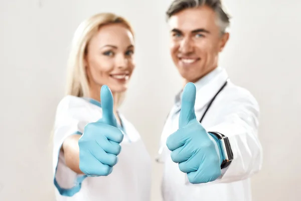 Two happy male and female doctors in medical uniform and blue gloves showing thumbs up, looking at camera and smiling, standing against grey background — Stock Photo, Image