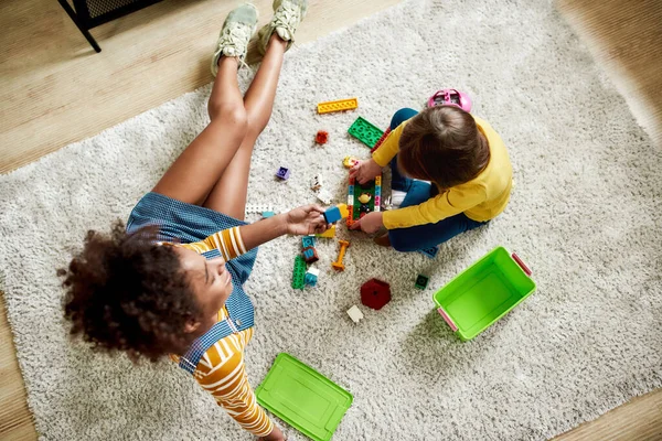Vista superior de niña caucásica pasar tiempo con niñera afroamericana, jugando con juguetes de construcción conjunto, sentado en el suelo — Foto de Stock