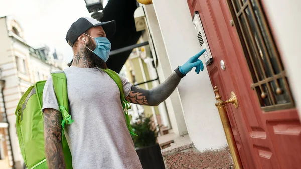 Brutal bearded delivery man in cap wearing mask due to the emergence of the Covid19 virus, ringing a doorbell, while delivering food — Stock Photo, Image