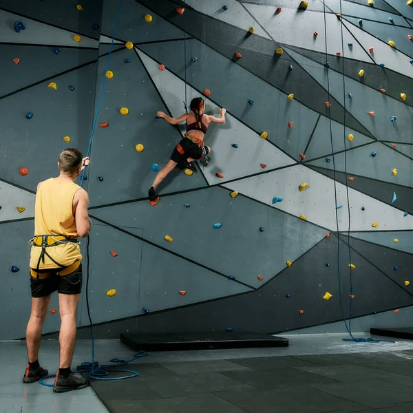 Instrutor masculino segurando corda, ensinando, olhando para a mulher em equipamentos de segurança e arnês enquanto ela está treinando na parede de escalada artificial — Fotografia de Stock