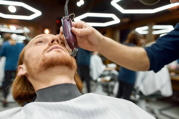 Поправка бороды. Крупным планом молодой красивый рыжий парень сидит в парикмахерском кресле и ухаживает за бородой в современной парикмахерской — стоковое фото