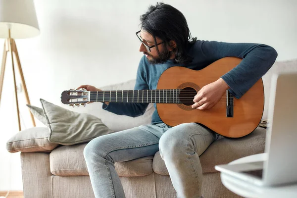 Learn and play. Young caucasian man sitting on sofa at home and adjusting acoustic guitar