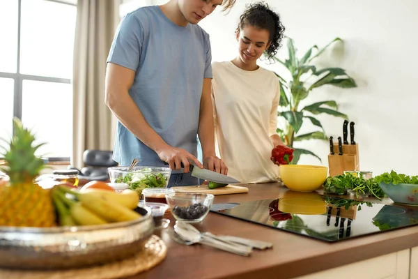 Νεαρός κόβει λαχανικά, ενώ γυναίκα κρατάει πιπέρι και τον παρακολουθεί. Χορτοφάγοι ετοιμάζουν υγιεινό γεύμα στην κουζίνα μαζί. Χορτοφαγία, υγιεινή διατροφή έννοια — Φωτογραφία Αρχείου