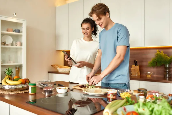Πιο εύχρηστος, πιο υγιής, πιο ευτυχισμένος. Νεαρός άνδρας κοπής λαχανικών, ενώ η γυναίκα έλεγχο συνταγή στην ψηφιακή ταμπλέτα. Χορτοφάγοι μαγειρεύουν πρωινό στην κουζίνα μαζί. Χορτοφαγία, υγιεινή διατροφή έννοια — Φωτογραφία Αρχείου