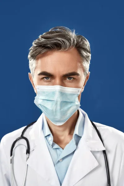 코로나 바이러스 그만 해. 의료용 마스크를 쓰고 푸른 해군 배경에 맞서 서서 카메라를 보고 있는 성숙 한 남성 의사의 실제 사진 — 스톡 사진