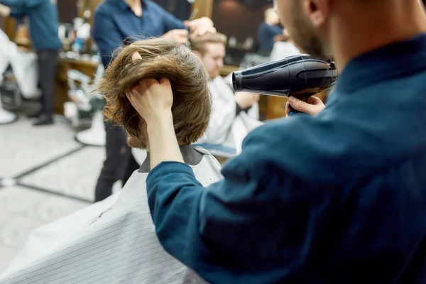 Back view of barber drying long hair of his client sitting in armchair in the modern barber shop — Stock Photo, Image