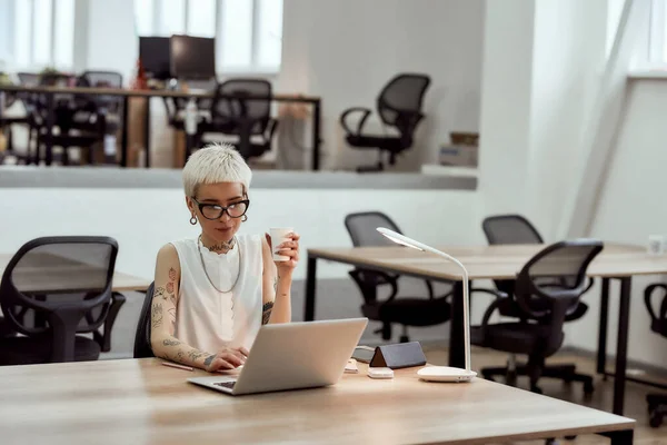 Nice coffee. Young concentrated tattooed business woman in eyewear holding cup of coffee and working with laptop while sitting alone in the modern office