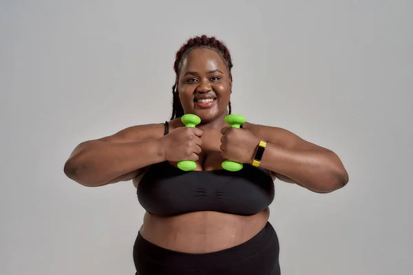 Plump, plus size afrikanisch amerikanische Frau in Sportbekleidung lächelnd, während des Trainings, mit grünen Hanteln im Studio vor grauem Hintergrund — Stockfoto