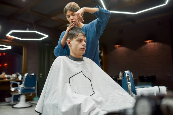 Jovem bonitão recebendo novo corte de cabelo moderno. Garota barbeiro profissional trabalhando com o cliente na barbearia ou salão de beleza. Concentre-se em jovem e cara — Fotografia de Stock