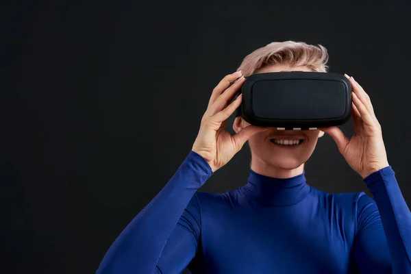Primer plano de mujer excitada con pelo corto en cuello alto azul con auriculares de realidad virtual, gafas vr aisladas sobre fondo oscuro —  Fotos de Stock