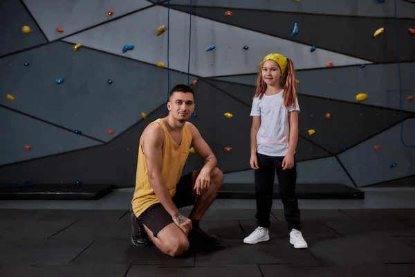 Erkek eğitmen ve aktif küçük kızın kameraya bakıp yapay tırmanma duvarına karşı durduğu uzun bir çekim. Spor hayatı ve kaya tırmanışı kavramı — Stok fotoğraf