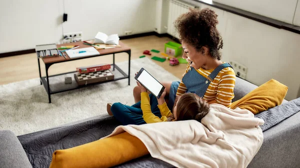 Pequena Maravilha. afro-americana baby sitter divertido caucasiano bonito menina. Kid está mostrando algo usando tablet pc, sentado no sofá — Fotografia de Stock