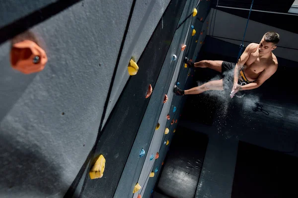 Junger Mann in Sicherheitsausrüstung und Gurtzeug trägt weißen Magnesiastaub auf, Kreide auf den Händen beim Training an der künstlichen Kletterwand drinnen — Stockfoto