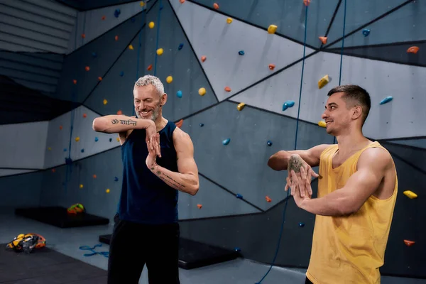 Joven instructor masculino y hombre de mediana edad sonriendo, calentando cuerpos, mientras se prepara para escalar en el centro de bouldering —  Fotos de Stock