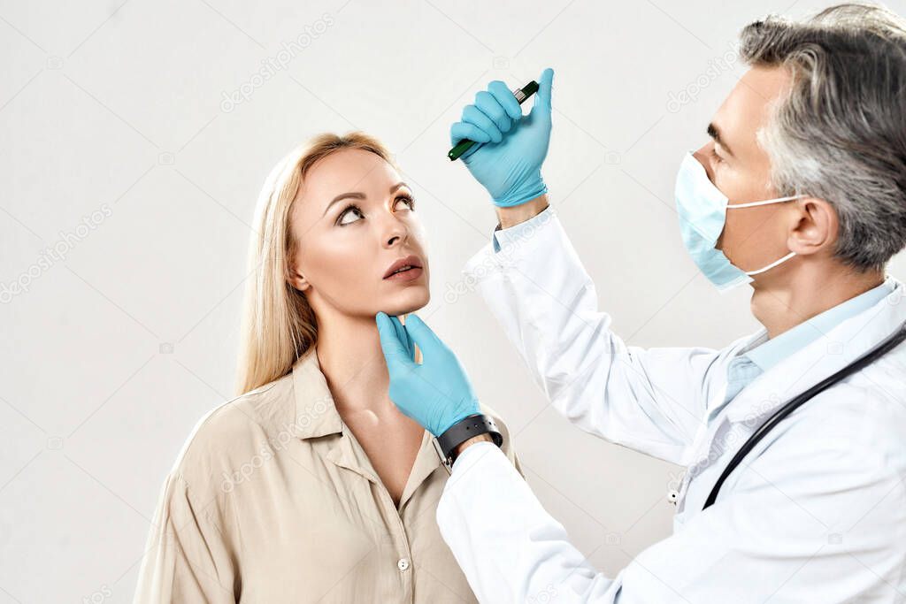 Professional mature male ophthalmologist in medical uniform examining eyes of young female patient while standing against grey background