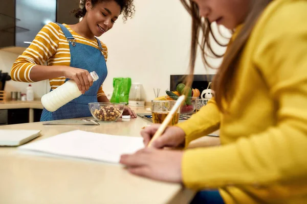 Alles, was ein Kind braucht. Afrikanische amerikanische Frau Babysitter unterhaltsam kaukasischen niedlichen kleinen Mädchen. Kind zeichnet, während Kindermädchen Mittagessen macht — Stockfoto