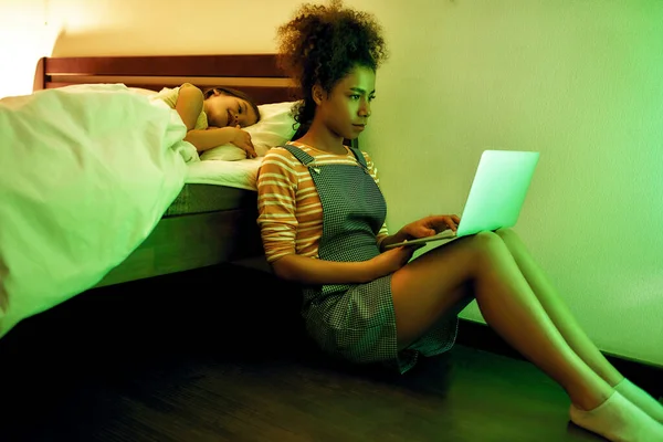 Atmósfera tranquila. Mujer afroamericana niñera pasando tiempo con una niña por la noche. Niñera usando laptop mientras niño mirándola y durmiendo en la cama —  Fotos de Stock
