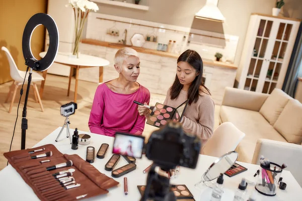 Helping you look good. Young female blogger, makeup artist applying makeup on middle aged woman face using cosmetic products while recording a tutorial video for her beauty blog