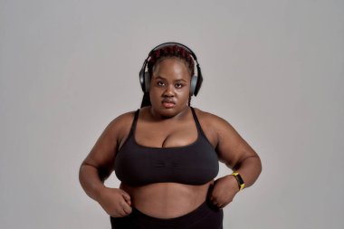 Vücuduna sahip çık. Şişkin, spor giyim ve kulaklık giyen Afro-Amerikan kadın kameraya bakıyor, gri arka planda stüdyoda duruyor. Spor anlayışı, sağlıklı yaşam tarzı, vücut pozitifi