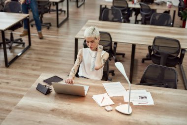 İş yerinde çalışan kadın bir ofis çalışanı. Ofiste otururken dizüstü bilgisayarıyla çalışan kısa saçlı, dövmeli genç bir iş kadını. İş ve kariyer