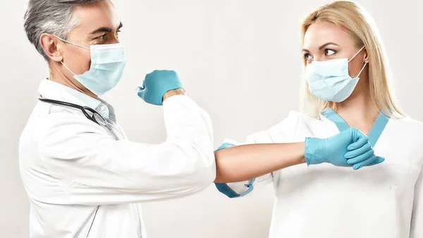 Social distancing. Two doctors in medical uniforms and protective masks greeting each other by bumping elbows, standing against grey background