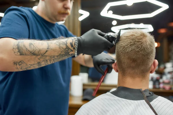 Barbiere professionista tatuato che lavora con tosatrice. Uomo in visita dal barbiere e ottenere un nuovo taglio di capelli moderno. Concentrati sulla testa di un cliente — Foto Stock