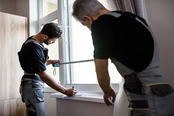Dos trabajadores profesionales en uniforme usando cinta métrica mientras miden la ventana y toman notas para instalar persianas en el interior. Concepto de construcción y mantenimiento —  Fotos de Stock