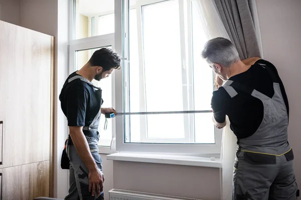 Два профессиональных работника в форме при помощи рулетки измеряют окно для установки жалюзи в помещении. Концепция строительства и обслуживания — стоковое фото