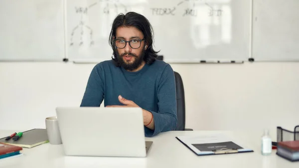 Учить программирование онлайн. Молодой целеустремленный бородатый учитель, преподающий информатику онлайн через видео-звонок, сидя на доске — стоковое фото