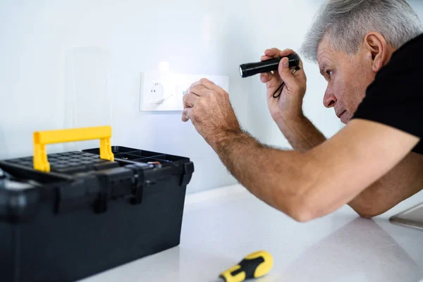 Пожилой электрик, ремонтник в форме работает, осматривает розетку на кухне при помощи фонарика. Концепция ремонта — стоковое фото